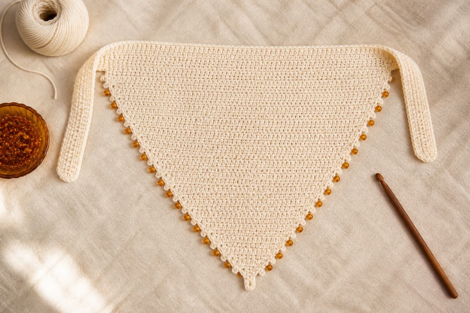 A cream crochet bandana with small amber beads along the sloping outer edges and long flat ties on linen.