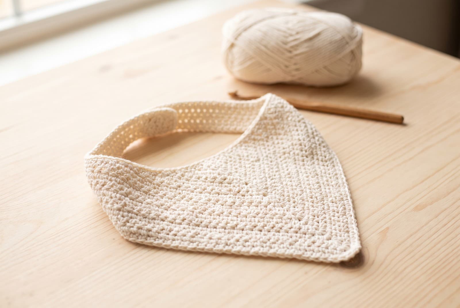 A cream crochet baby bandana bib laid flat on a wooden surface beside a ball of cotton yarn and a wooden hook
