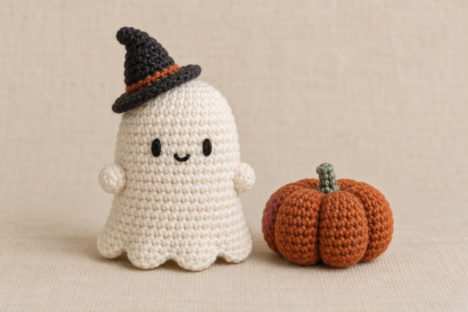 Ivory crocheted ghost with embroidered eyes, tiny bobble arms and scalloped hem, wearing a charcoal witch hat beside an orange pumpkin.